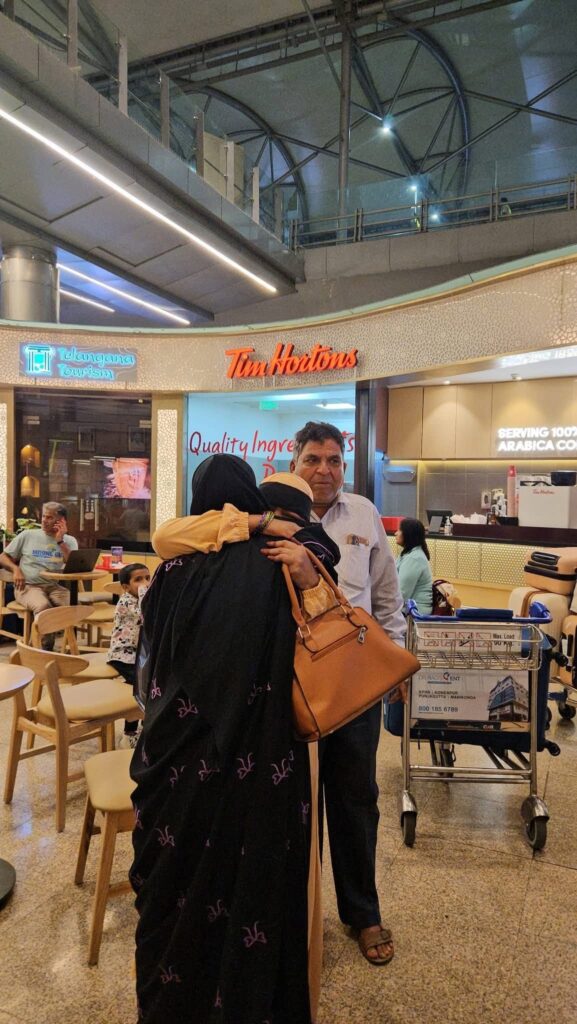 A woman in a black abaya hugs a family member at an airport, with luggage and seating visible in the background.