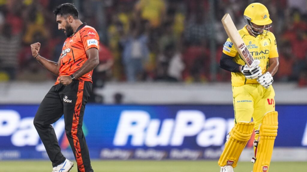 Two cricket players, one in orange and one in yellow, celebrating during a match at a stadium.