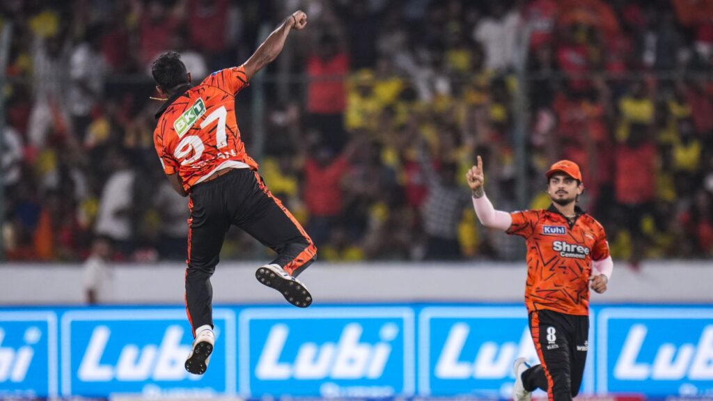 Two cricket players in orange uniforms celebrating a wicket in a stadium with cheering crowd.