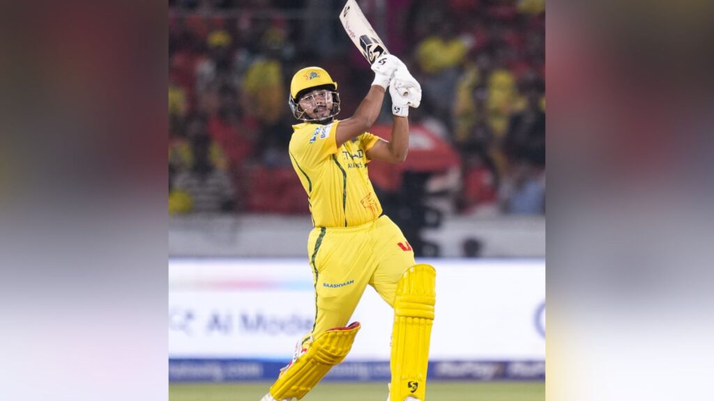 Cricket player in yellow uniform swinging bat during match, focused on ball, with crowd in background.