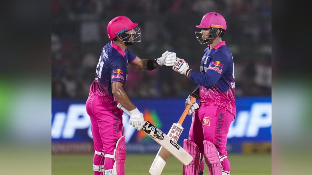 Two female cricket players in pink uniforms exchanging a fist bump after a successful play.