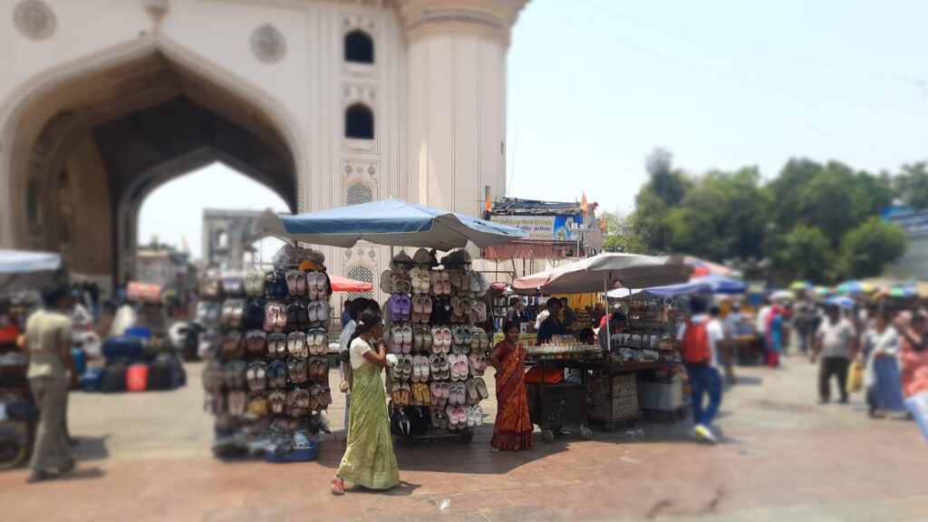 Street market stall selling hats and accessories near historic monument in India, busy with shoppers.