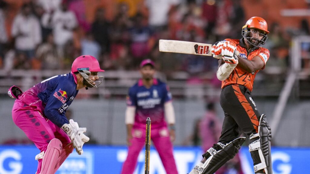 A cricket player in orange and black gear batting during a match, with a wicketkeeper in pink ready behin.