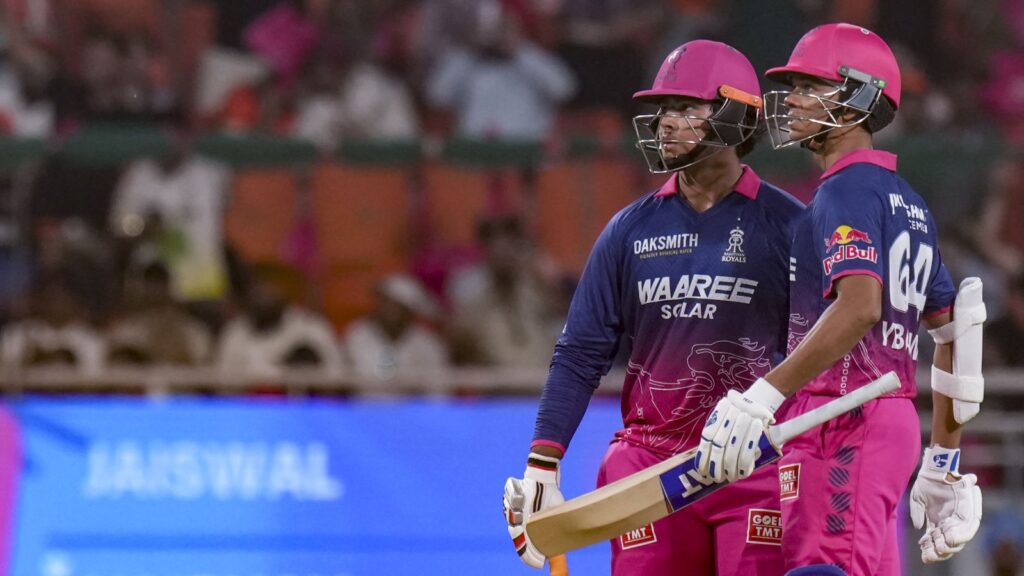 Two female cricket players in pink and blue uniforms holding a bat during a match at a stadium.